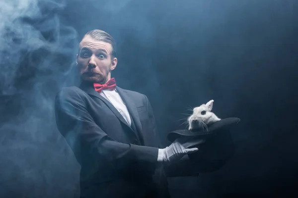 Emocionalmente sorprendido mago sosteniendo conejo blanco en sombrero, cuarto oscuro con humo - foto de stock