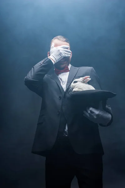 Mágico emocional fechar os olhos e mostrando truque com coelho branco no chapéu, sala escura com fumaça — Fotografia de Stock