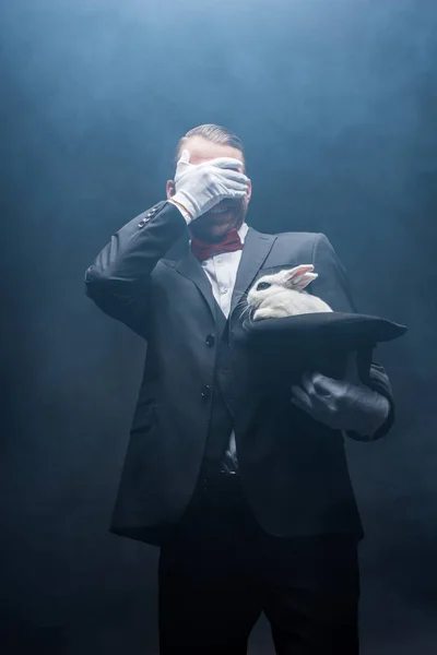 Emotional magician closing eyes and showing trick with white rabbit in hat, dark room with smoke — Stock Photo