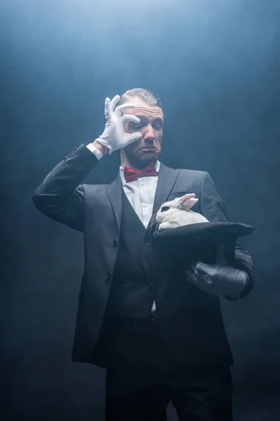 Mago sorprendido mirando conejo blanco en sombrero, cuarto oscuro con humo - foto de stock