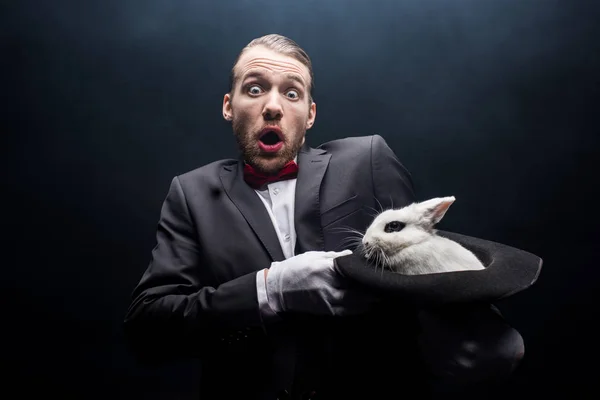 Shocked magician holding white rabbit in hat, dark room with smoke — Stock Photo