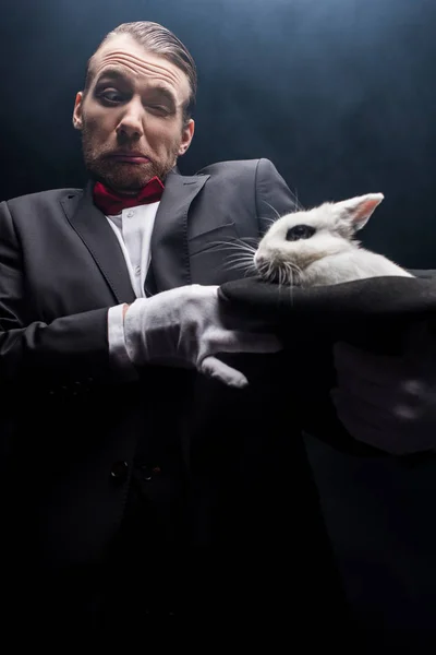 Заплутаний фокусник тримає білого кролика в капелюсі, темна кімната з димом — стокове фото