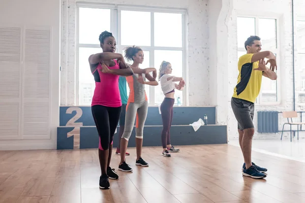 Vista lateral do belo treinador praticando movimentos de zumba com dançarinos multiétnicos no estúdio de dança — Fotografia de Stock