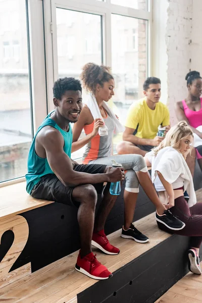 Focus sélectif de l'homme afro-américain avec bouteille de sport souriant à la caméra par des danseurs multiculturels sur banc en studio de danse — Photo de stock