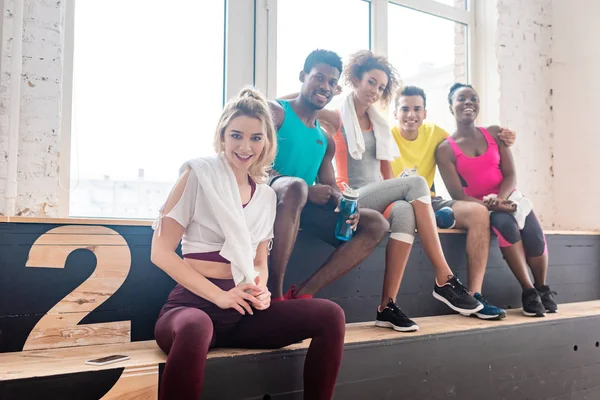 Selective focus of multicultural zumba dancers smiling at camera on bench in dance studio — Stock Photo