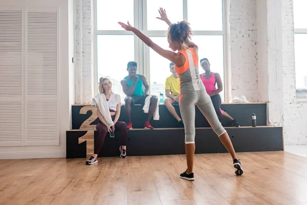 Enfoque selectivo de la mujer realizando movimientos con bailarinas de zumba multiculturales descansando en banco en estudio de danza - foto de stock