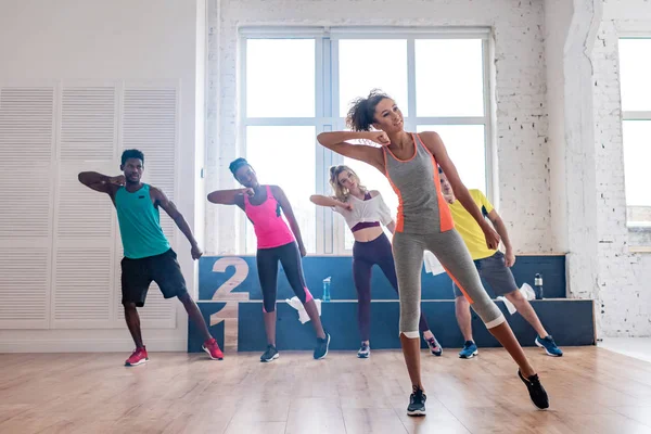 Entraîneur afro-américain souriant jouant de la zumba avec des danseurs multiculturels dans un studio de danse — Photo de stock