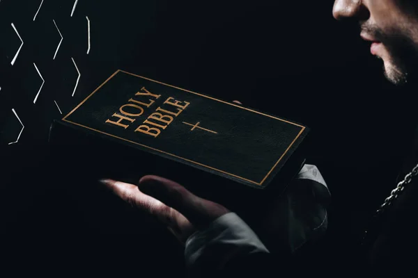 Vista recortada del sacerdote católico sosteniendo la sagrada Biblia cerca de la rejilla confesional en la oscuridad con rayos de luz - foto de stock