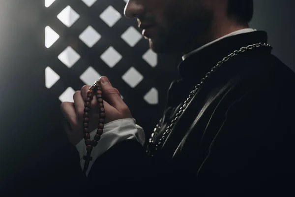 Teilansicht des katholischen Priesters mit hölzernen Rosenkranzperlen in der Nähe des Beichtstuhlgitters im Dunkeln mit Lichtstrahlen — Stockfoto