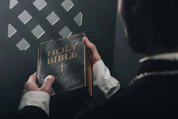 Cropped view of catholic priest holding holy bible near confessional grille in dark with rays of light — Stock Photo