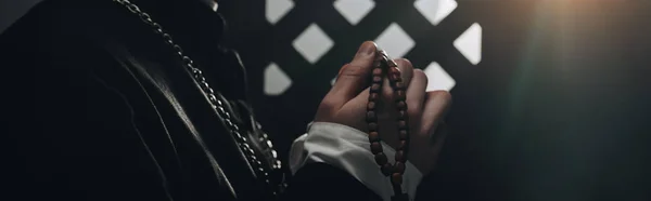 Vista parcial del sacerdote católico sosteniendo cuentas de rosario de madera cerca de la rejilla confesional en la oscuridad con rayos de luz, plano panorámico - foto de stock
