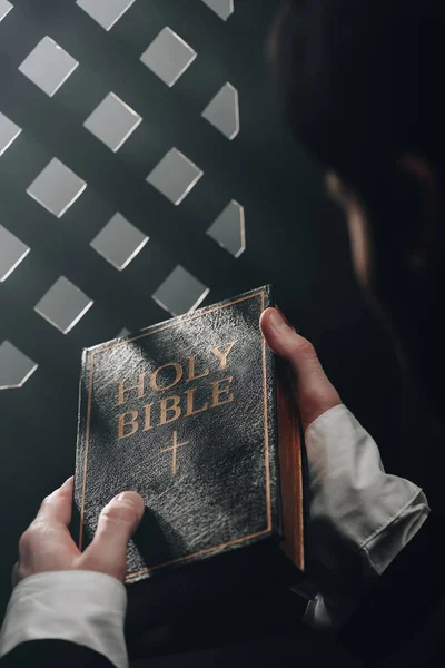 Vista parcial del sacerdote católico sosteniendo la sagrada Biblia cerca de la rejilla confesional en la oscuridad con rayos de luz - foto de stock