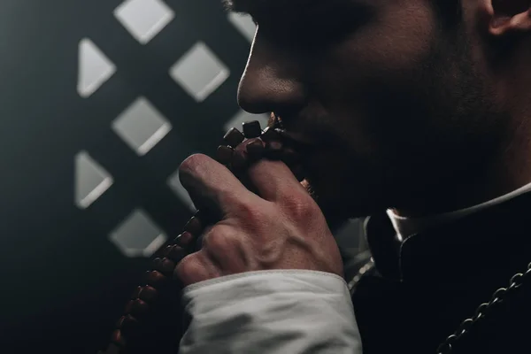 Young catholic priest kissing wooden rosary beads near confessional grille in dark with rays of light — Stock Photo