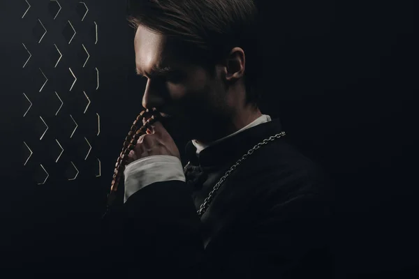 Young thoughtful catholic priest kissing wooden rosary beads near confessional grille in dark with rays of light — Stock Photo