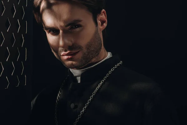 Young confident catholic priest looking at camera near confessional grille in dark with rays of light — Stock Photo