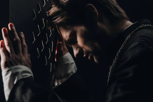 Young tense catholic priest with closed eyes leaning on confessional grille in dark with rays of light — Stock Photo