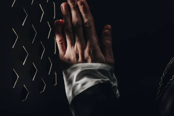 Teilansicht eines katholischen Priesters, der in der Nähe eines Beichtstuhlgitters im Dunkeln mit Lichtstrahlen betet — Stockfoto
