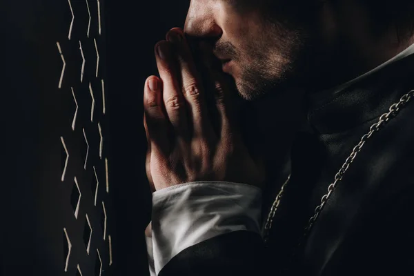 Обрезанный вид католического священника молящегося рядом с исповедальней решеткой в темноте с лучами света — стоковое фото