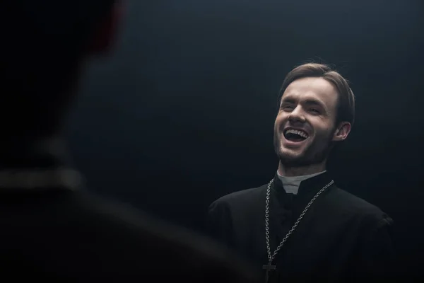 Young catholic priest laughing sarcastically while looking at own reflection isolated on black — Stock Photo