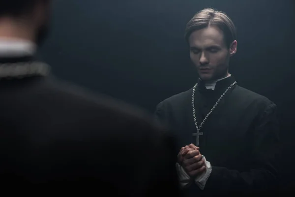 Young serious catholic priest praying with closed eyes near own reflection isolated on black — Stock Photo