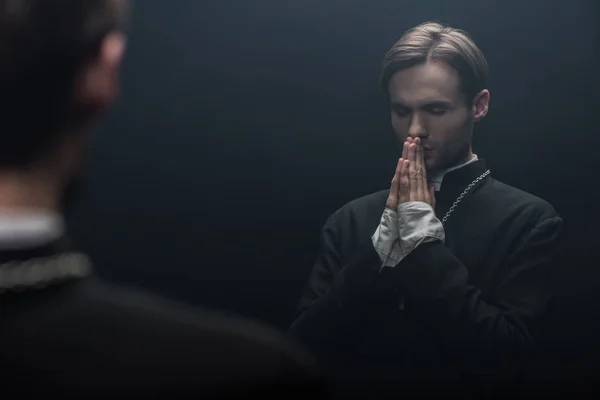 Young serious catholic priest praying with closed eyes near own reflection isolated on black — Stock Photo