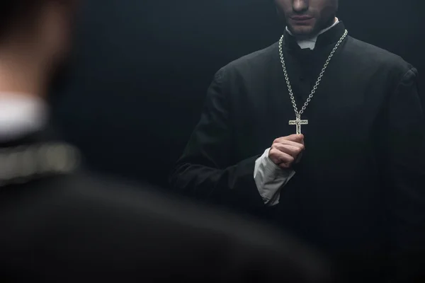 Abgeschnittene Ansicht des katholischen Priesters, der das Kreuz an seiner Halskette berührt, in der Nähe seines eigenen Spiegels isoliert auf schwarz — Stockfoto