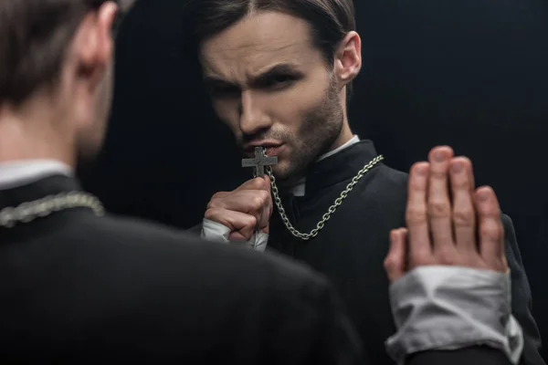 Young tense catholic priest kissing holy cross while looking at own reflection isolated on black — Stock Photo