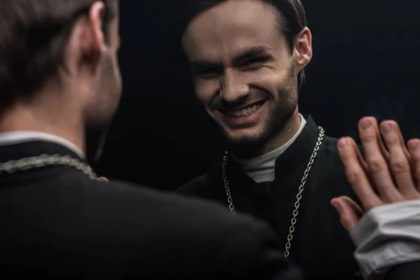 Молодий католицький священик сміється саркастично, дивлячись на власне відображення ізольовано на чорному — стокове фото