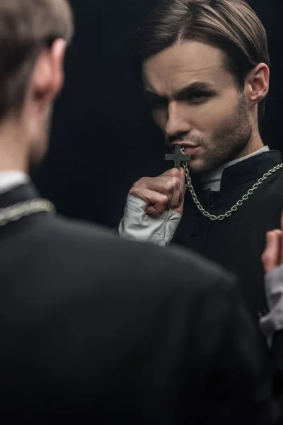 Young tense catholic priest kissing holy cross while looking at own reflection isolated on black — Stock Photo