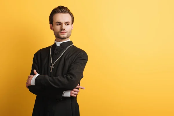 Sicuro sacerdote cattolico in piedi con le braccia incrociate e distogliendo lo sguardo isolato sul giallo — Foto stock