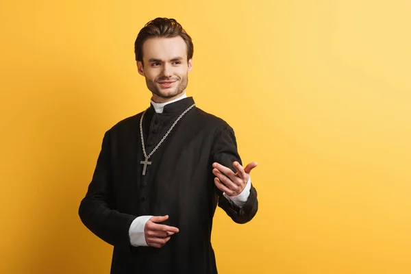 Sonriente sacerdote católico de pie con la mano extendida mientras mira hacia otro lado aislado en amarillo - foto de stock