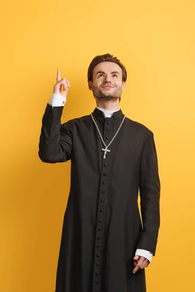 Sonriente sacerdote católico mirando hacia arriba y señalando con el dedo aislado en amarillo - foto de stock