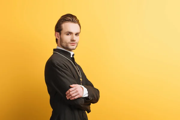 Confident catholic priest with crossed arms looking at camera isolated on yellow — Stock Photo