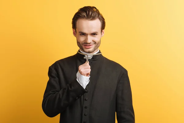 Young catholic priest holding cross on his necklace while smiling isolated on yellow — Stock Photo
