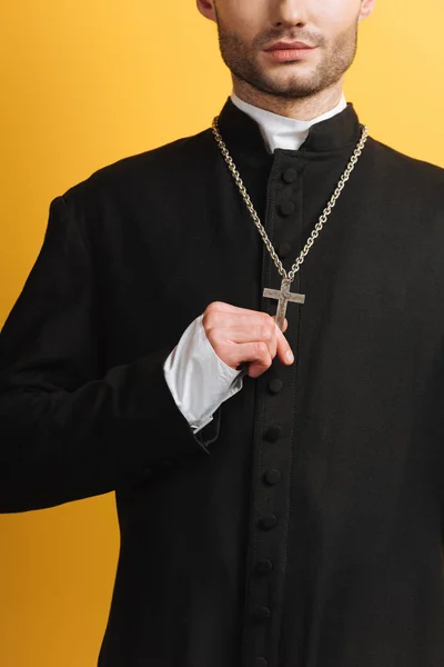 Vista parcial del sacerdote católico tocando la cruz en su collar aislado en amarillo - foto de stock