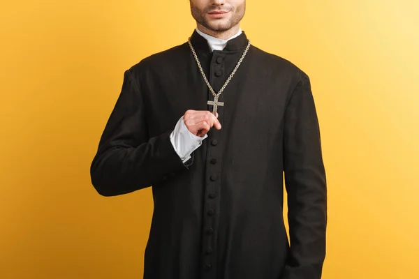 Обрізаний вид на католицького священика, який торкається хреста на його намисті ізольовано на жовтому — стокове фото