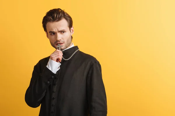 Sacerdote católico tenso mirando hacia otro lado mientras sostiene la cruz cerca de los labios aislados en amarillo - foto de stock