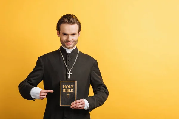 Sonriente sacerdote católico señalando con el dedo la sagrada biblia mientras mira a la cámara aislada en amarillo - foto de stock