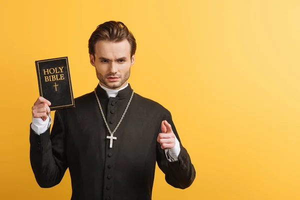Serious catholic priest pointing with finger at camera while holding bible isolated on yellow — Stock Photo