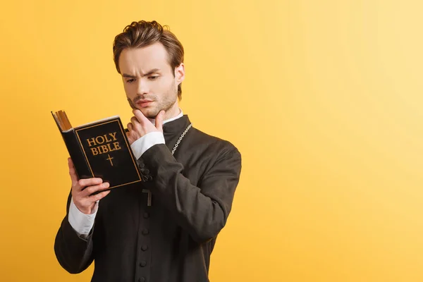 Concentrated catholic priest touching face while reading bible isolated on yellow — Stock Photo