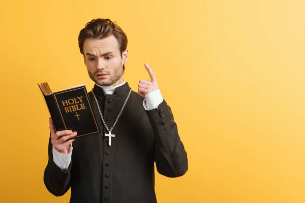 Pensativo sacerdote católico mostrando signo de idea mientras que la lectura de la Biblia aislado en amarillo - foto de stock