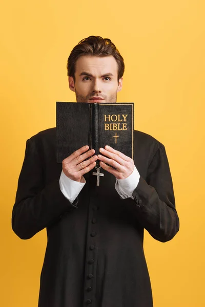 Thoughtful catholic priest looking up while holding open bible isolated on yellow — Stock Photo