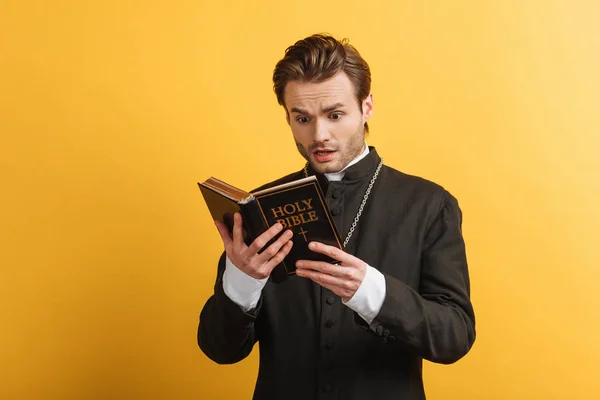 Shocked catholic priest reading holy bible isolated on yellow — Stock Photo