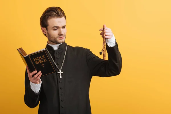 Thoughtful catholic priest holding bible and looking at wooden rosary beads isolated on yellow — Stock Photo