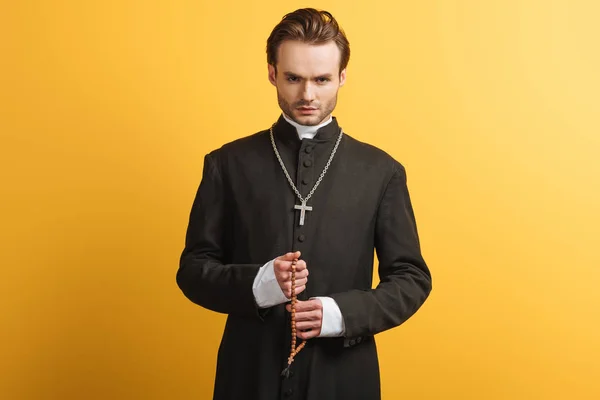 Молодий впевнений католицький священик тримає дерев'яні бісеру ізольовані на жовтому — стокове фото