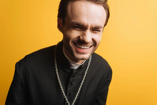 Збуджений католицький священик сміється, дивлячись на камеру ізольовано на жовтому — стокове фото