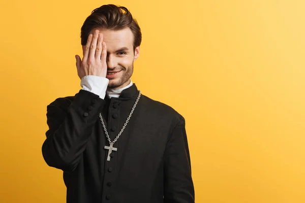 Sonriente sacerdote católico cubriendo el ojo con la mano mientras mira a la cámara aislada en amarillo - foto de stock