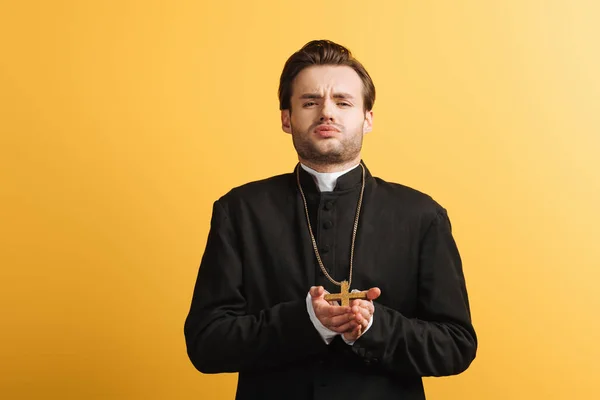 Young catholic priest with bored face expression holding cross and looking at camera isolated on yellow — Stock Photo