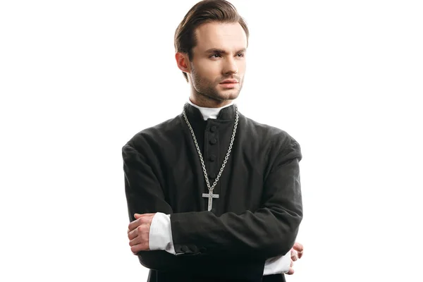 Joven y confiado sacerdote católico mirando hacia otro lado mientras está de pie con los brazos cruzados aislados en blanco - foto de stock