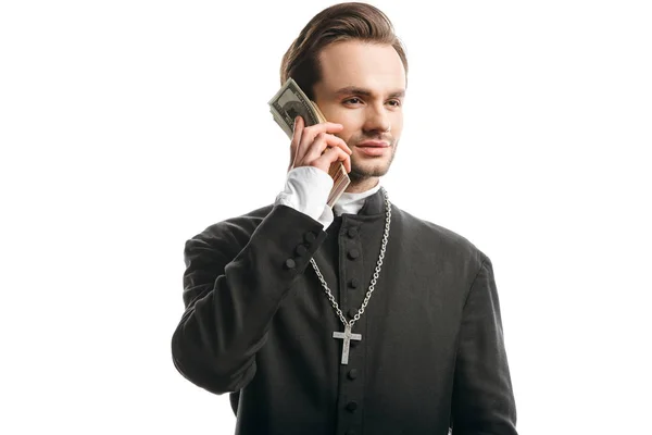Sacerdote católico corrupto mirando hacia otro lado mientras sostiene el dinero cerca de la cara aislado en blanco - foto de stock